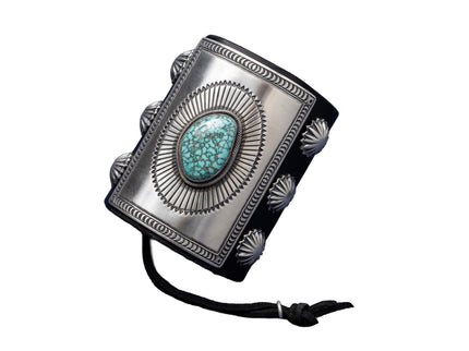 A bold leather bowguard adorned with a rectangular sterling silver faceplate, finely stamped in a sunburst motif framing a sky-blue Kingman turquoise stone with dark matrix. Embellished with fan-pattern silver conchos on both sides and tied with a black leather cord.