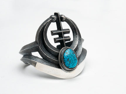 A silver bracelet with a turquoise centerpiece, featuring a geometric motif and oxidized silver elements.