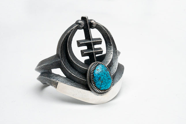 A silver bracelet with a turquoise centerpiece, featuring a geometric motif and oxidized silver elements.