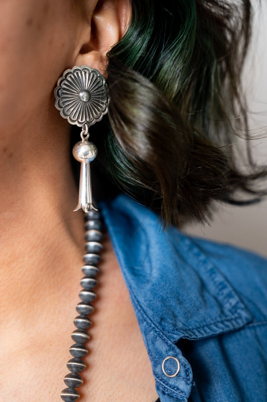 Close-up of a circular silver drop earring with a silver tassel hanging from the ear of a Navajo woman wearing a blue shirt.