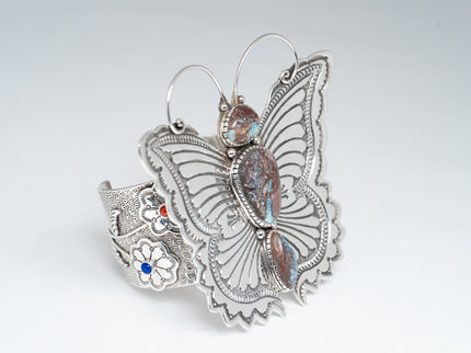 Angled front of bracelet showcasing a silver stamped butterfly with antennas and three turquoise stones. Side Cuff displays two flowers with one coral and one lapis stone.