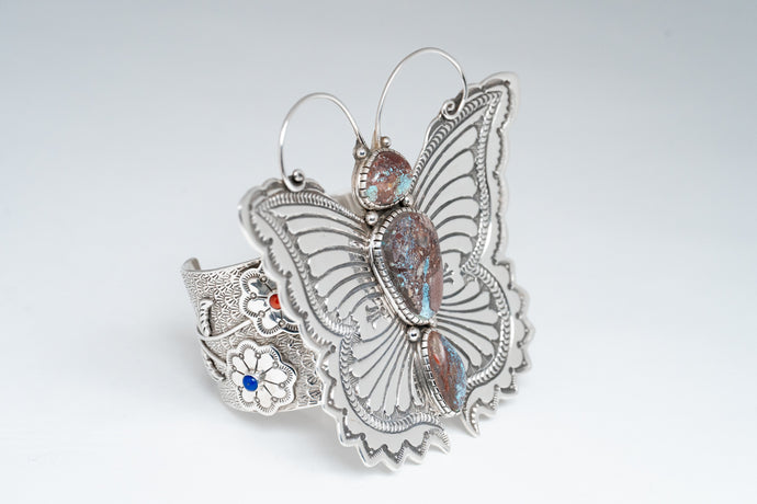 Angled front of bracelet showcasing a silver stamped butterfly with antennas and three turquoise stones. Side Cuff displays two flowers with one coral and one lapis stone.