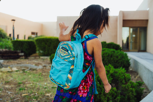Little Navajo girl wearing a colorful dress and blue Pendleton backpack in front of a college building
