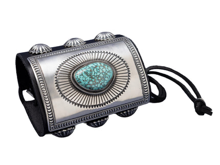 A bold leather bowguard adorned with a rectangular sterling silver faceplate, finely stamped in a sunburst motif framing a sky-blue Kingman turquoise stone with dark matrix. Embellished with fan-pattern silver conchos on both sides and tied with a black leather cord.