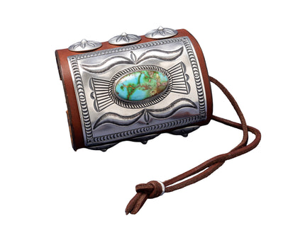 A richly detailed leather bowguard with a rectangular sterling silver plate showcasing flowing stamped patterns and a striking green-blue Sonoran Gold turquoise stone at the center. Decorated with dome-shaped silver conchos and secured with a dark brown leather tie.