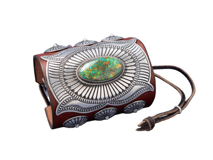 A handcrafted leather bowguard featuring an elaborate stamped sterling silver overlay with a radiant sunburst design, centered around a vibrant Sonoran Gold turquoise cabochon. Accented with scalloped conchos on the sides and a brown leather strap for fastening.