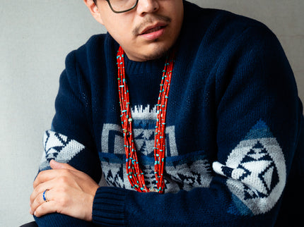Navajo man wearing a dark blue Pendleton sweater with geometric patterns and a red coral necklace against a gray background