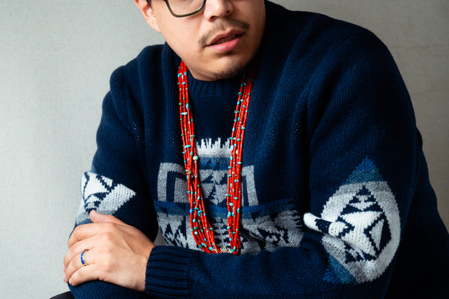 Navajo man wearing a dark blue Pendleton sweater with geometric patterns and a red coral necklace against a gray background