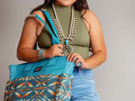 Young Navajo woman wearing blue jeans and a OD green top smiling and carrying a Pendleton blue tote bag on her right shoulder. She is standing in front of a beige background and wearing a squash blossom and large turquoise bracelet
