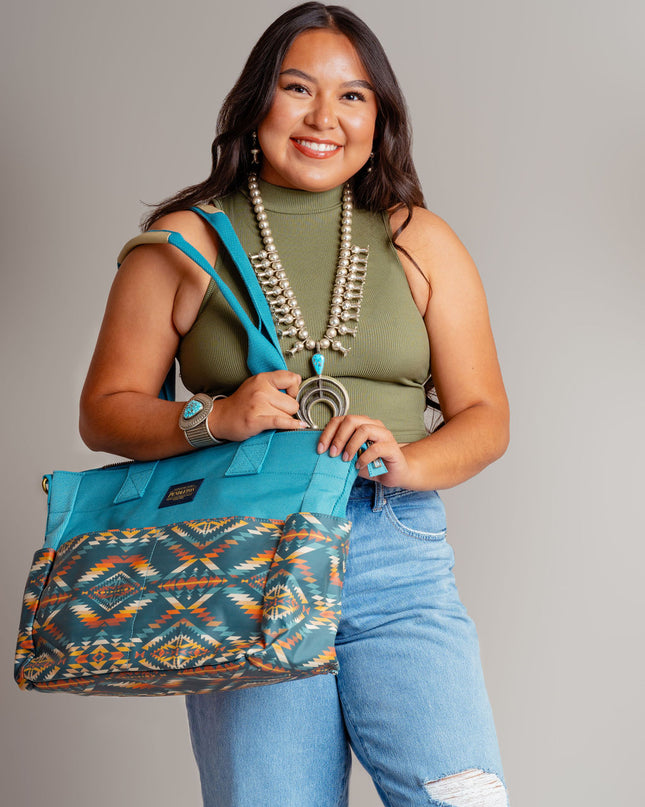 Young Navajo woman wearing blue jeans and a OD green top smiling and carrying a Pendleton blue tote bag on her right shoulder. She is standing in front of a beige background and wearing a squash blossom and large turquoise bracelet