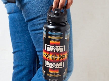 Person holding a water bottle with a colorful pattern, wearing blue jeans and an orange turquoise bracelet.