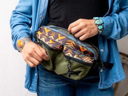 Person wearing a blue button up shirt with blue jeans is unzipping a Pendleton patterned fanny pack they are wearing