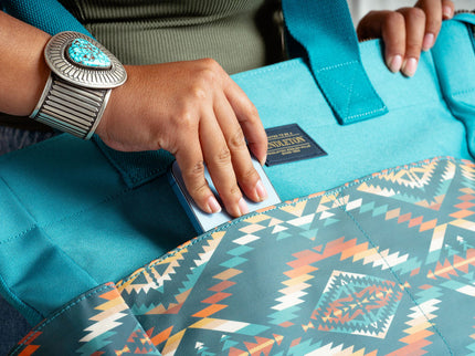 Close up of a females's right hand placing a blue smart phone into the outside pocket  of a Pendleton tote bag. Model is wearing a large turquoise bracelet on her wrist.