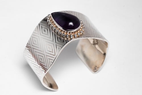 A silver bracelet with diamond-shaped stamped patterns and a large deep purple sugalite stone, featuring a chain of smaller beads.