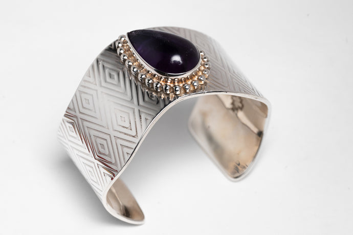 A silver bracelet with diamond-shaped stamped patterns and a large deep purple sugalite stone, featuring a chain of smaller beads.