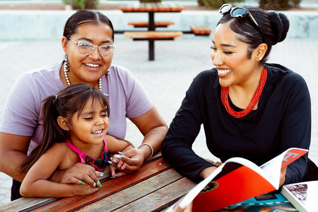 Three Navajo women sitting at a picnic table outdoors, reading a book together.