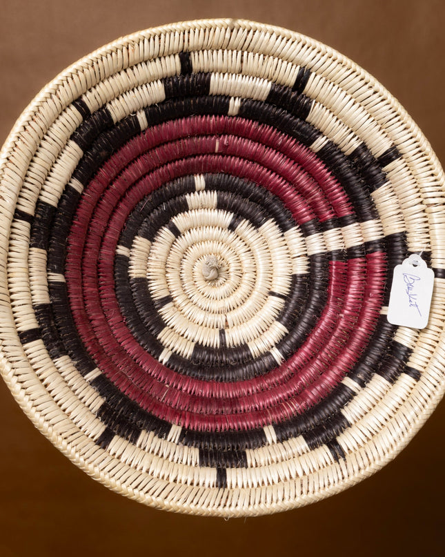 A vintage Navajo basket with a starburst pattern in natural, red, and black tones, shown against a warm brown background.