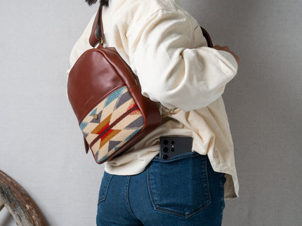 Navajo woman wearing a brown leather backpack with a geometric woven pattern, with a phone in back pants pocket, against a creme background.
