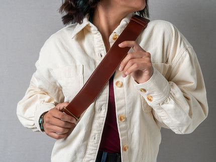 Person wearing a creme colored button shirts holds the brown leather belt strap of a small backpack 