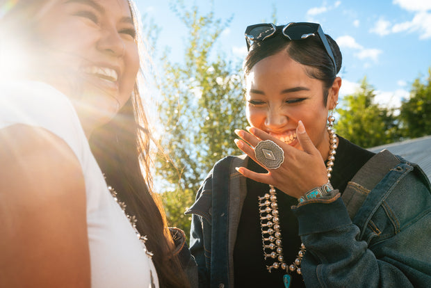 Two Navajo women laughing outdoors with trees and blue sky in the background and wearing Navajo jewelry with modern clothing