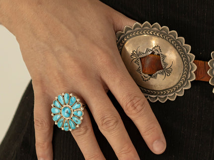 A close-up of a woman's hand wearing a large silver ring with turquoise stones, resting against a black ribbed garment and a Navajo silver concho belt.