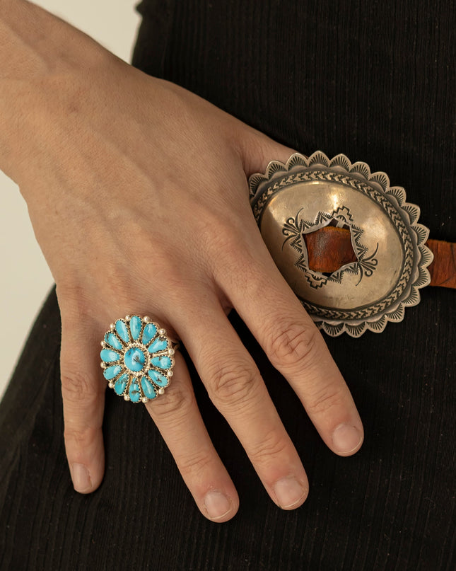 A close-up of a woman's hand wearing a large silver ring with turquoise stones, resting against a black ribbed garment and a Navajo silver concho belt.