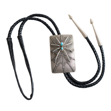 Full view of a bolo tie that has the design of silver spider with two turquoise stones as the body raised on a rectangular silver Bolo tie with spider web etching with a black leather cord and two silver tips