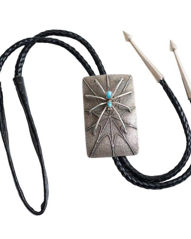 Full view of a bolo tie that has the design of silver spider with two turquoise stones as the body raised on a rectangular silver Bolo tie with spider web etching with a black leather cord and two silver tips