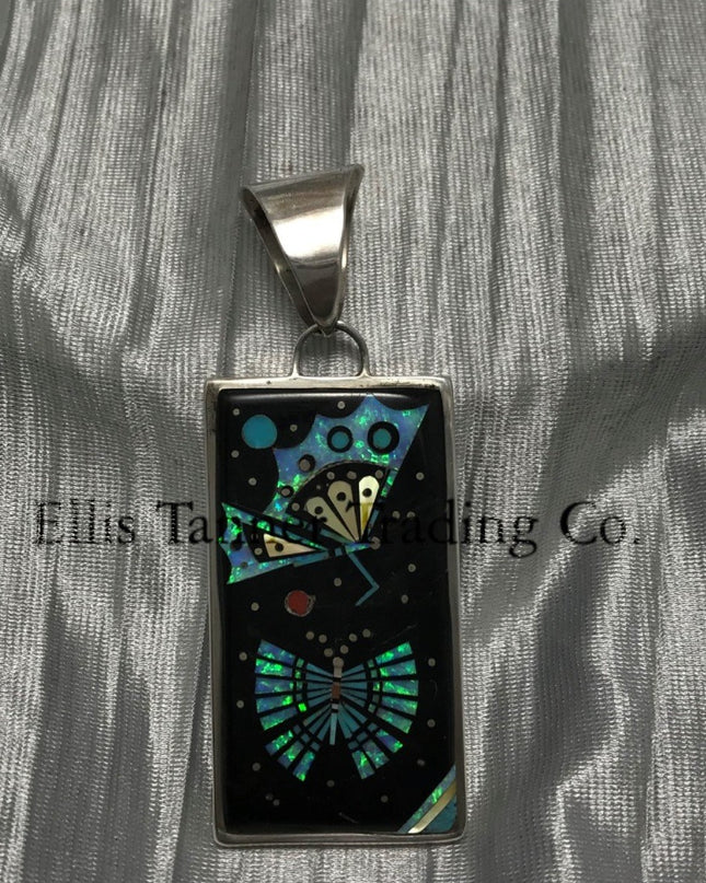 A rectangular pendant with a silver frame and loop for a chain. The pendant features an intricate design with geometric patterns in vibrant turquoise, black, and yellow, resembling a butterfly and spreading it wings. The background has shimmering details, creating a striking contrast.