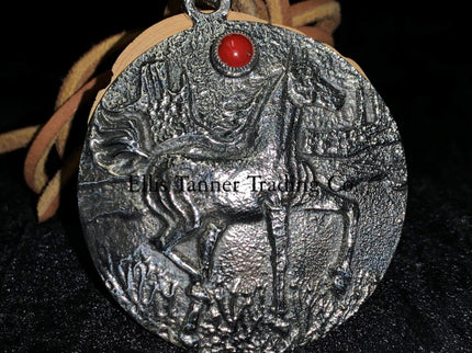 A circle silver pendant with a sand cast horse and one coral stone, with a tan leather strap. Displayed on top of a black cloth 