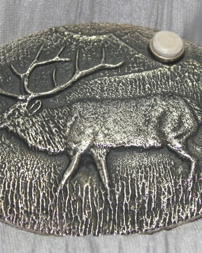 A sand-cast buckle featuring an intricately etched elk design set against a textured background. Added is a small, antler-shaped stone, adding a distinctive and natural touch. The highlighting its rustic and artisanal craftsmanship.