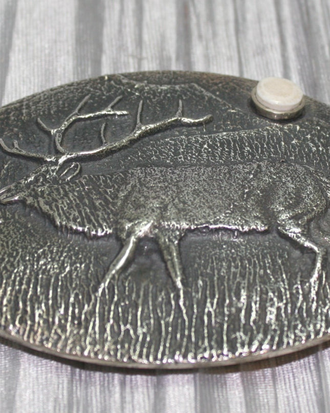 A sand-cast buckle featuring an intricately etched elk design set against a textured background. Added is a small, antler-shaped stone, adding a distinctive and natural touch. The highlighting its rustic and artisanal craftsmanship.