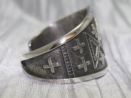 handcrafted silver cuff bracelet featuring intricate geometric and cross patterns etched into the metal. The design is bold and symmetrical, with a textured background that enhances the detailed craftsmanship. The bracelet rests on a neutral surface, emphasizing its artistic and traditional appeal.