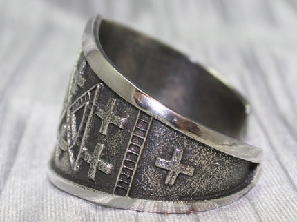 handcrafted silver cuff bracelet featuring intricate geometric and cross patterns etched into the metal. The design is bold and symmetrical, with a textured background that enhances the detailed craftsmanship. The bracelet rests on a neutral surface, emphasizing its artistic and traditional appeal.