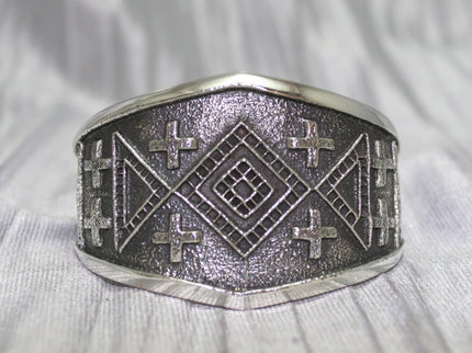 handcrafted silver cuff bracelet featuring intricate geometric and cross patterns etched into the metal. The design is bold and symmetrical, with a textured background that enhances the detailed craftsmanship. The bracelet rests on a neutral surface, emphasizing its artistic and traditional appeal.