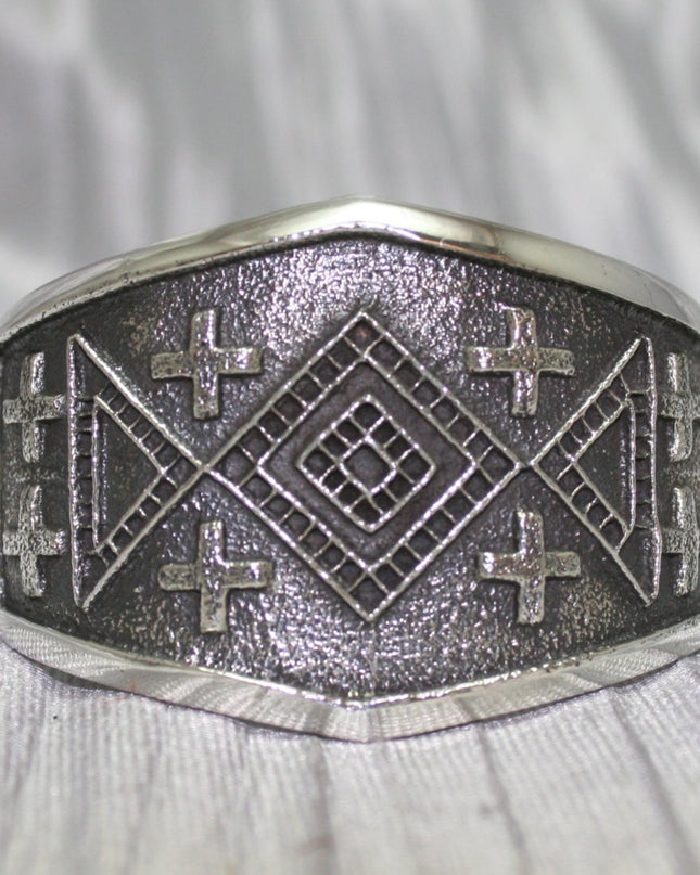 handcrafted silver cuff bracelet featuring intricate geometric and cross patterns etched into the metal. The design is bold and symmetrical, with a textured background that enhances the detailed craftsmanship. The bracelet rests on a neutral surface, emphasizing its artistic and traditional appeal.