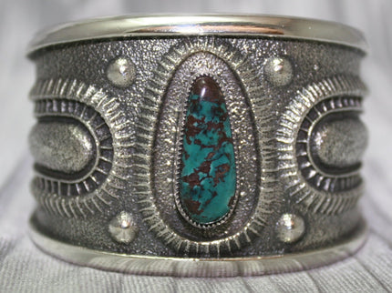A bold sand-cast cuff featuring intricate textures and patterns, with a vibrant oval Pilot Mountain turquoise stone at the center. The turquoise showcases deep blue tones with a coppery matrix, framed by an ornate border that adds depth and elegance. The design reflects a balance of rugged craftsmanship and refined artistry.