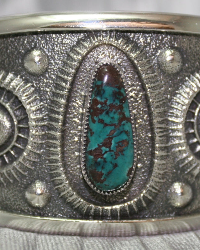 A bold sand-cast cuff featuring intricate textures and patterns, with a vibrant oval Pilot Mountain turquoise stone at the center. The turquoise showcases deep blue tones with a coppery matrix, framed by an ornate border that adds depth and elegance. The design reflects a balance of rugged craftsmanship and refined artistry.