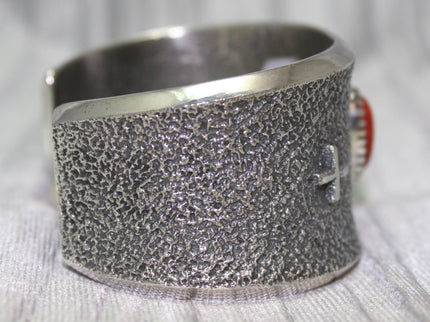 A wide silver cuff bracelet with a textured surface, small silver cross on side. The cuff rests on a soft gray background, highlighting its intricate craftsmanship.