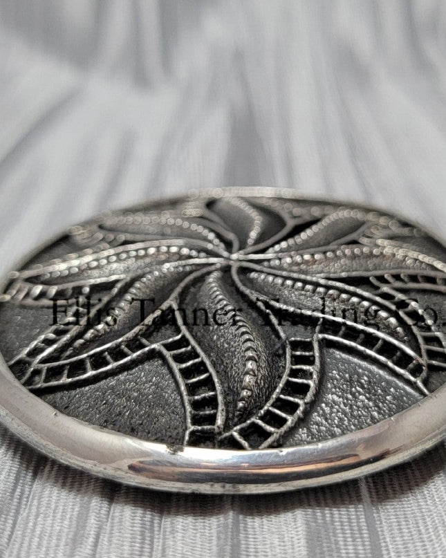 Silver sandcast, spiral flower, buckle 
 side view, placed on top of a silver cloth.