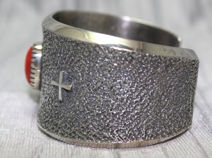 A wide silver cuff bracelet with a textured surface, small silver cross on side. The cuff rests on a soft gray background, highlighting its intricate craftsmanship.