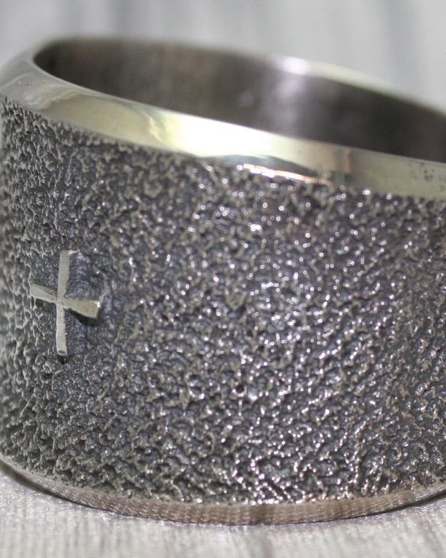 A wide silver cuff bracelet with a textured surface, small silver cross on side. The cuff rests on a soft gray background, highlighting its intricate craftsmanship.