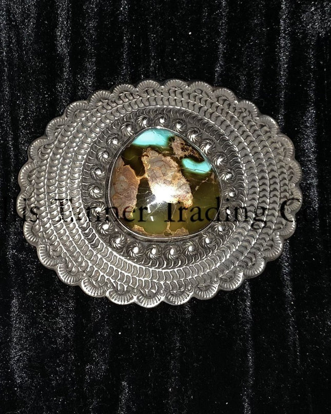 A silver belt buckle with a Royston turquoise centerpiece and intricate button details on a black background.