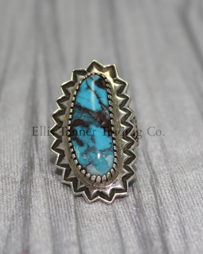 An oval-shaped Bisbee Turquoise with black vein matrix. surrounded in a silver starburst border. this ring is displayed on top of a gray cloth 