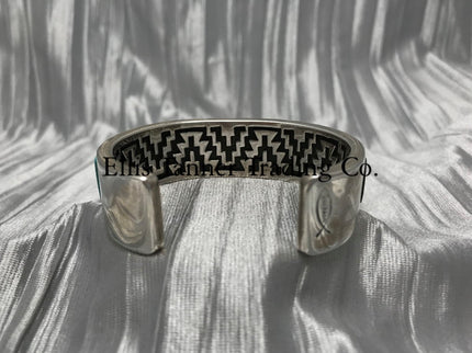 Displays the inside of the bracelet features a geometric zigzag pattern, resembling a traditional or tribal design, engraved into the metal. The pattern alternates sharply, creating a bold visual contrast against the smooth outer surface.