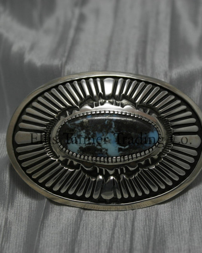 Oval shaped buckle, with a center Black diamond turquoise stone, displayed on top of a shiny silver sheet 