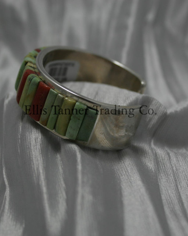 A silver bracelet featuring an intricate cobblestone inlay design with vibrant Carico Lake turquoise and coral stones. The piece is displayed on a soft, textured silver-gray fabric, side profile 
