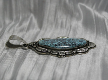A handcrafted sterling silver pendant with a Morenci Turquoise gemstone. The stone is blue with a black and gray matrix and is set in an ornate silver frame with scalloped and stamped detailing. The pendant features a large bail for easy attachment to a chain.