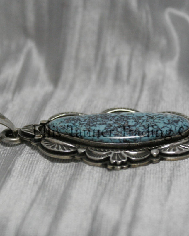 A handcrafted sterling silver pendant with a Morenci Turquoise gemstone. The stone is blue with a black and gray matrix and is set in an ornate silver frame with scalloped and stamped detailing. The pendant features a large bail for easy attachment to a chain.