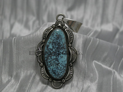 A handcrafted sterling silver pendant with a Morenci Turquoise gemstone. The stone is blue with a black and gray matrix and is set in an ornate silver frame with scalloped and stamped detailing. The pendant features a large bail for easy attachment to a chain.